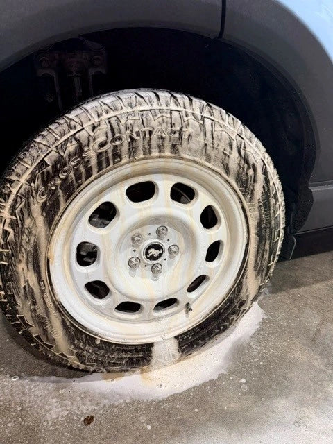 A close-up of a car wheel covered in thick white soap foam, sitting on a concrete floor with a puddle of soapy water in front. The tire tread is heavily foamed, and the silver metal wheel is visible.
